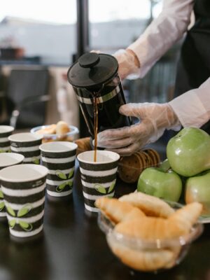 Barista pours coffee from French press into cups at a breakfast event with croissants and apples.